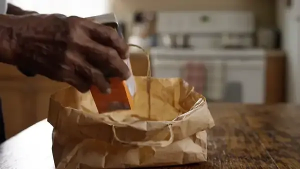 Hands packing a small carton and two amber bottles into a brown paper bag at a kitchen table